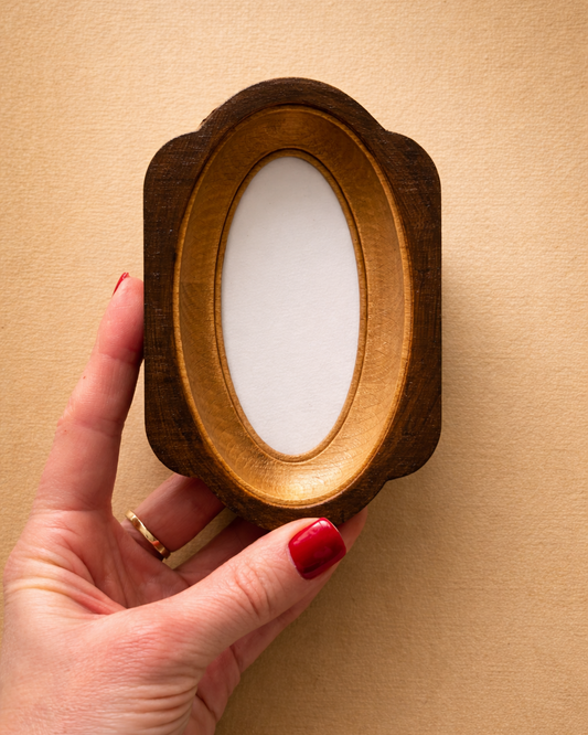 Wooden oval frame held by a hand with red nail polish against a beige background