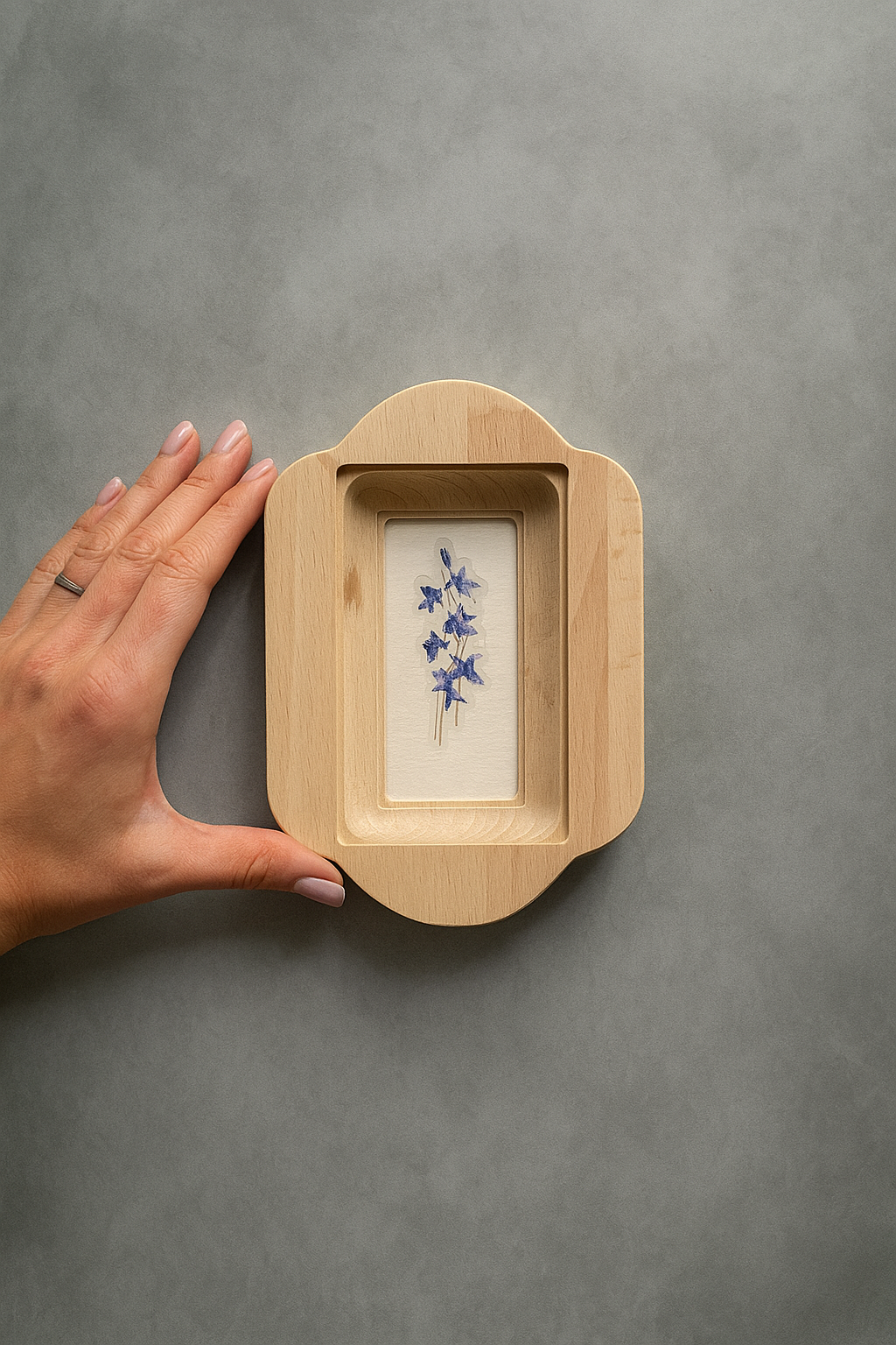 Hand holding a wooden frame with a small white card inside containing blue flowers on a gray background