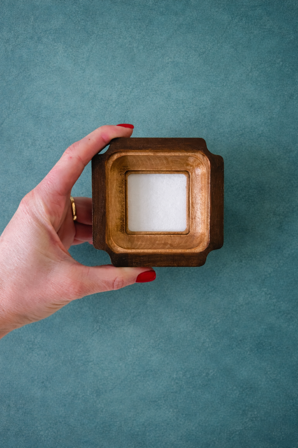 Hand holding a wooden square frame with a white square inside against a teal background
