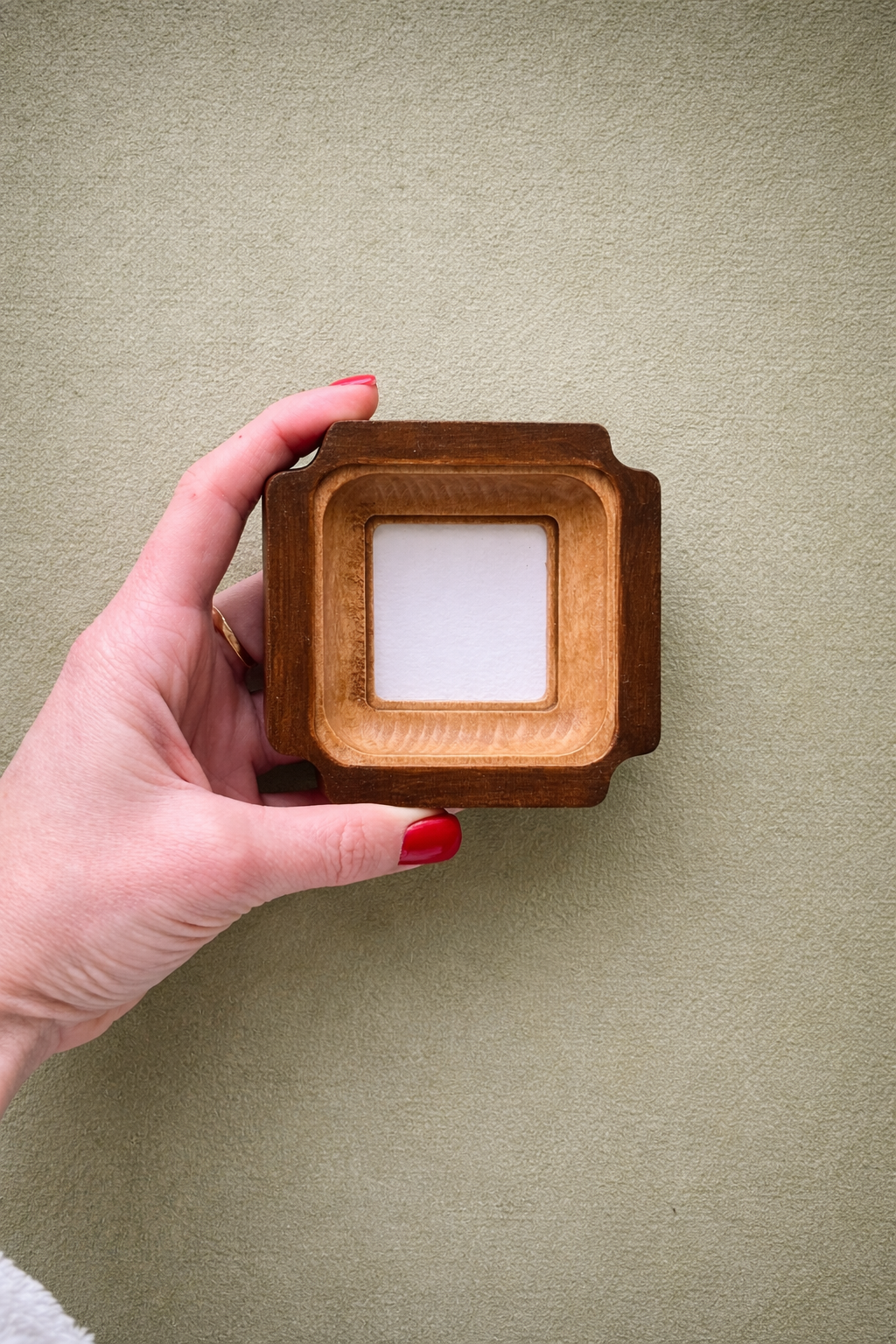 Hand holding a wooden picture frame with a white square inside against a beige background